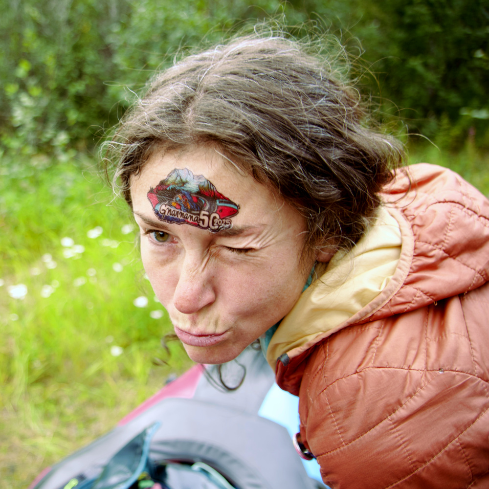A woman making a goofy face with the Gnarnana logo tattooed on her forehead. She's wearing a light red puffy jacket and a blurred packraft and vegetation is visible in the background.