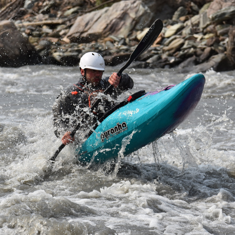 A kayaker catching air off of a wave in a gray glacial river. The kayak is blue and purple and there are water droplets spraying in the air around the kayaker.