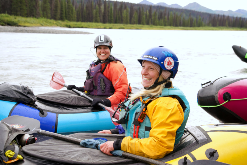 two packrafters on the Nenana River with spruce trees in the background and an ochre cliff on the right with a brilliant blue sky above