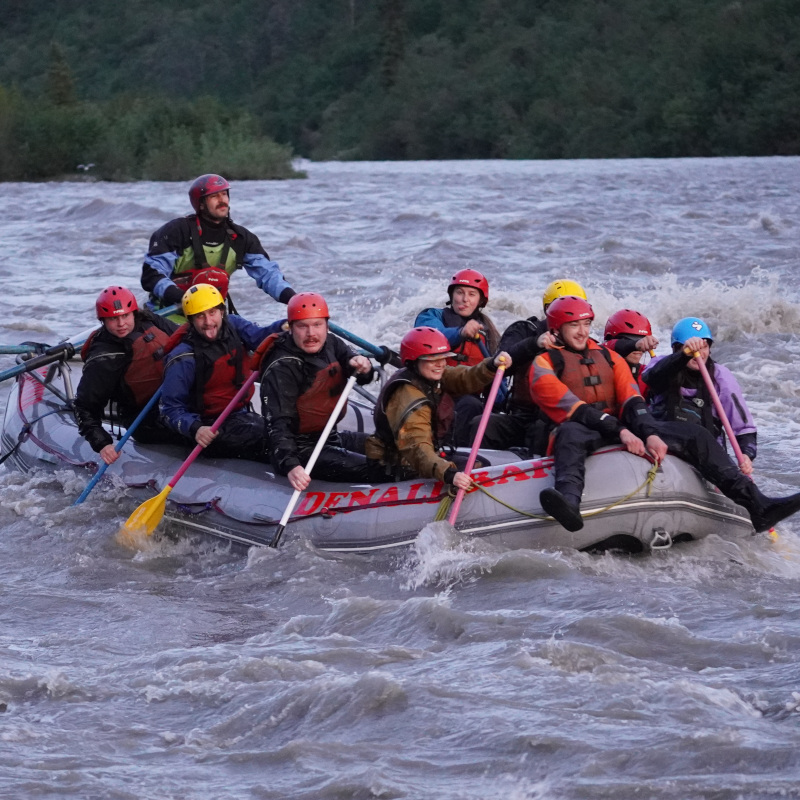 A raft full of people making funny faces at Two Rocks rapids on the Nenana River in the evening with alpenglow. The raft is gray and the river is a glacial gray color. PFDs, drysuits, and helmets are all bright and varied colors.