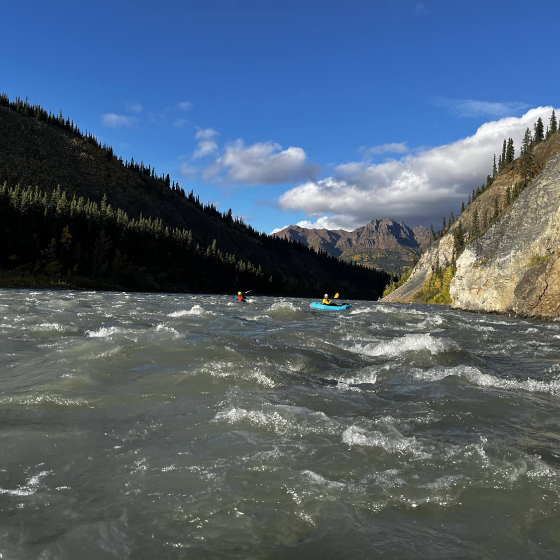 two packrafters on the Nenana River with Mount Fellows in the background and an ochre cliff on the right with a brilliant blue sky above