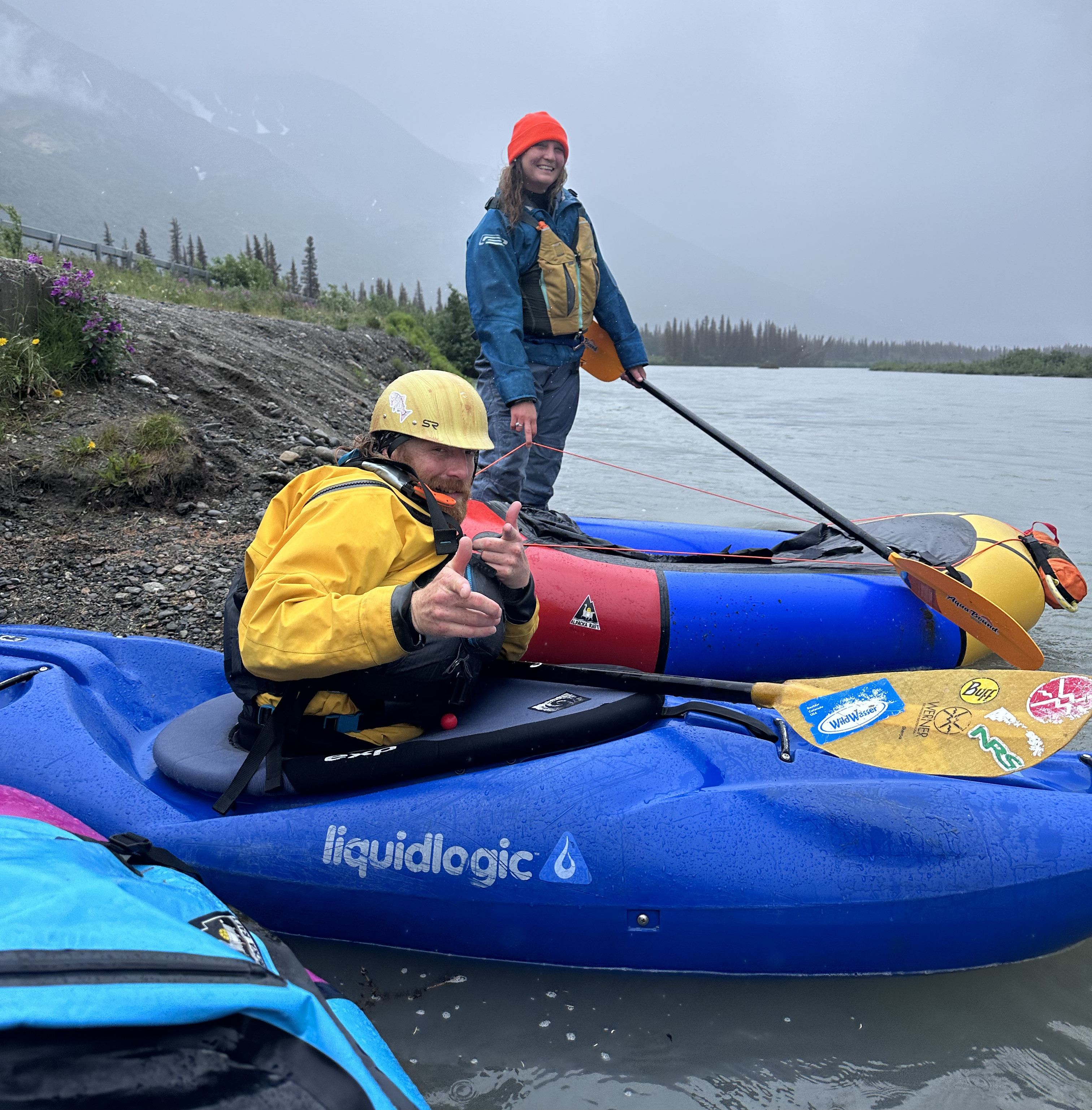A kayaker and a packrafter looking at the camera as they get ready to put in on a large glacial river. The kayaker is giving finger guns to the camera.